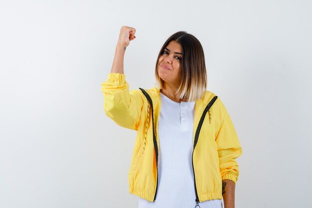 Молодая девушка показывает жест победителя в белой футболке, желтой куртке и выглядит удачливым, вид спереди.