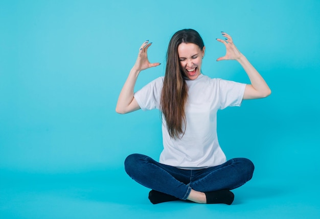 Young girl si screaming by raising up her hands and sitting on floor on blue background