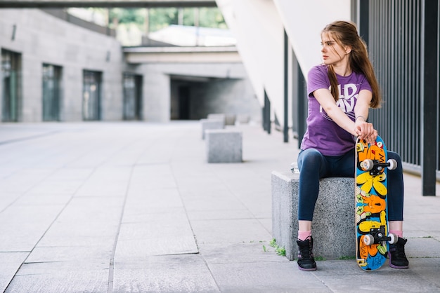 若い女の子は、カラフルなスケートボードを右に見て座っている