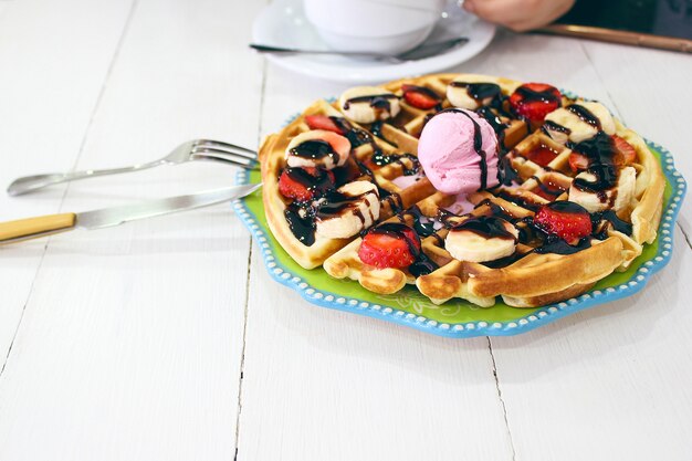 Free photo young girl sitting caffe eating breakfast waffle with chocolate sauce, banana slices and strawberries on green ceramic plate and photographed her breakfast