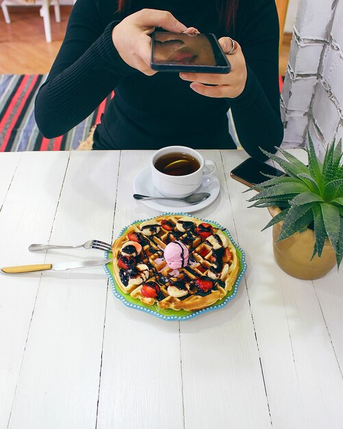 緑のセラミックプレートにチョコレートソース、バナナのスライス、イチゴの朝食ワッフルを食べるカフェに座っている若い女の子と彼女の朝食を撮影