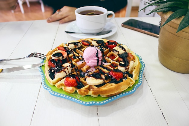 緑のセラミックプレートにチョコレートソース、バナナのスライス、イチゴの朝食ワッフルを食べるカフェに座っている若い女の子と彼女の朝食を撮影