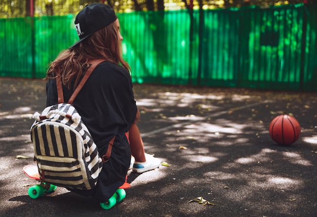 Young girl sitting on plastic orange penny shortboard on asphalt in cap