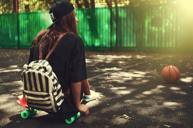 Young girl sitting on plastic orange penny shortboard on asphalt in cap