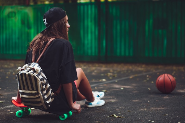 Молодая девушка сидит на пластиковой оранжевой копеечке на асфальте в кепке