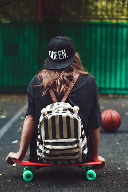 Young girl sitting on plastic orange penny shortboard on asphalt in cap