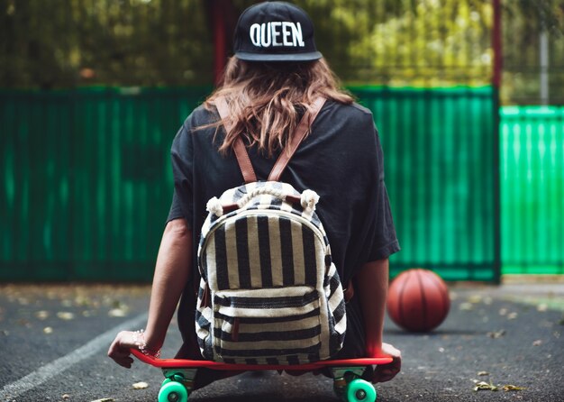 Young girl sitting on plastic orange penny shortboard on asphalt in cap