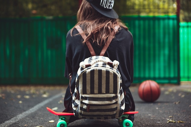 Young girl sitting on plastic orange penny shortboard on asphalt in cap