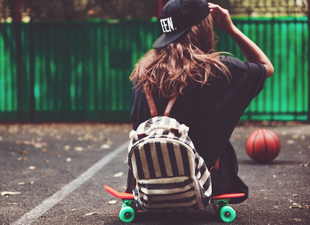 Young girl sitting on plastic orange penny shortboard on asphalt in cap