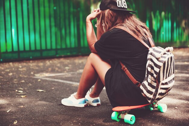 Young girl sitting on plastic orange penny shortboard on asphalt in cap