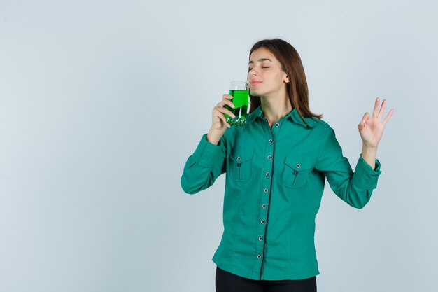 Ragazza giovane odorando un bicchiere di liquido verde, mostrando segno ok in camicetta verde, pantaloni neri e guardando sanguigno, vista frontale.