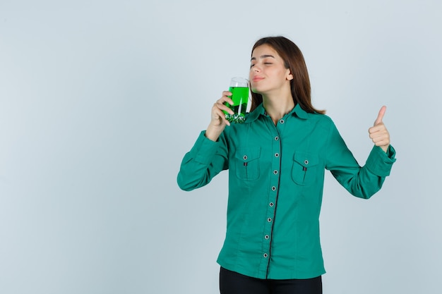 Young girl smelling glass of green liquid, showing thumb up in green blouse, black pants and looking sanguine , front view.