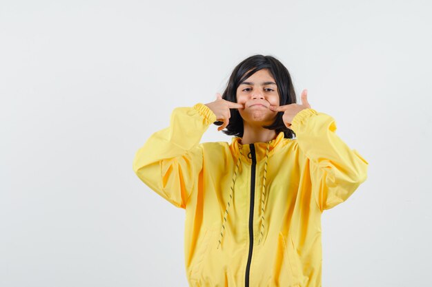 Young girl smiling and pointing index finger to cheeks in yellow bomber jacket and looking serious.