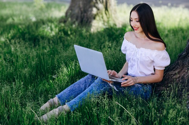 Молодая девушка улыбается, сидя на траве с ноутбуком на ее ноги