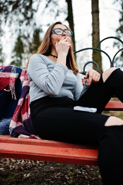 Молодая девушка курит сигарету на открытом воздухе, сидя на скамейке Концепция никотиновой зависимости подростков