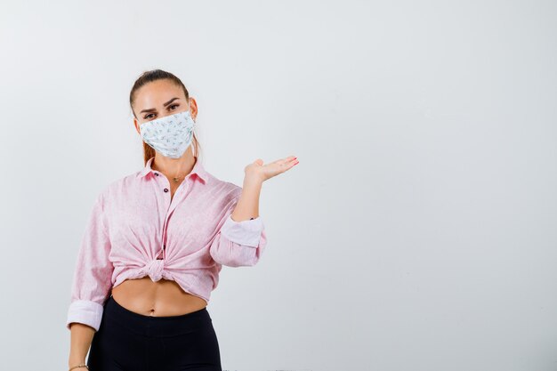 Young girl spreading palm aside in pink blouse, black pants, mask and looking alluring. front view.