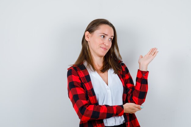 Young girl spreading palm in checkered shirt, blouse and looking curious , front view.