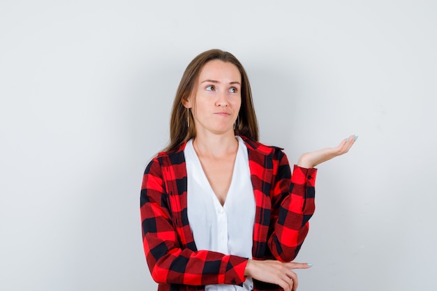 Young girl spreading palm in checkered shirt, blouse and looking thoughtful , front view.