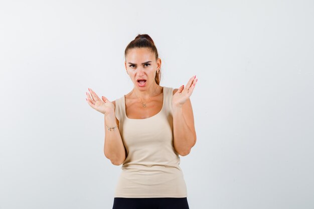 Young girl spreading palms out, keeping mouth wide open in beige top, black pants and looking surprised , front view.