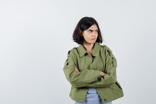 Young girl standing arms crossed, looking away in grey sweater, khaki jacket, jean pant and looking focused. front view.