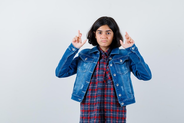 체크 셔츠와 진 재킷을 입고 서 있는 어린 소녀가 진지한 표정으로 손가락을 건넜습니다.