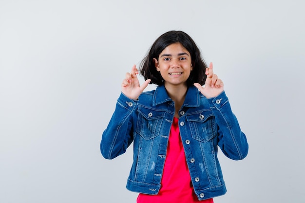 Young girl standing fingers crossed in red t-shirt and jean jacket and looking happy. front view.