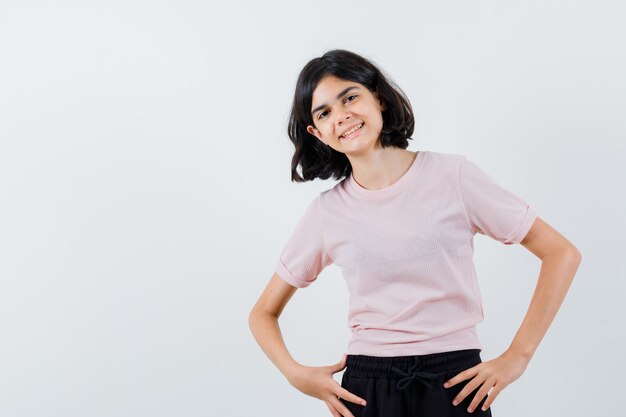 Young girl standing straight, holding hands on waist and posing at camera in pink t-shirt and black pants and looking happy