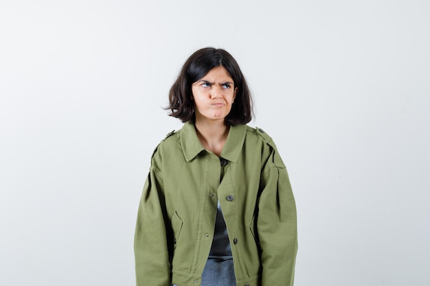 Young girl standing straight, looking away and posing  in grey sweater, khaki jacket, jean pant and looking annoyed , front view.