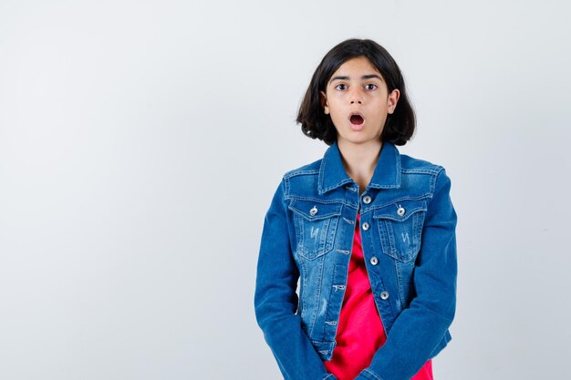 Young girl standing straight, opening mouth and posing at camera in red t-shirt and jean jacket and looking surprised , front view.