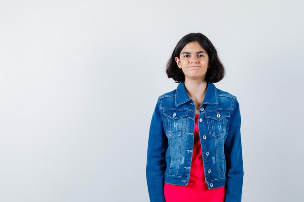 Young girl standing straight and posing at camera in red t-shirt and jean jacket and looking happy. front view.