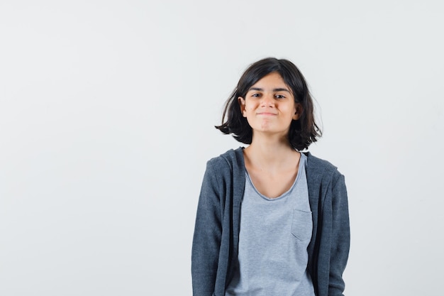 Young girl standing straight, smiling and posing at camera in light gray t-shirt and dark grey zip-front hoodie and looking cute