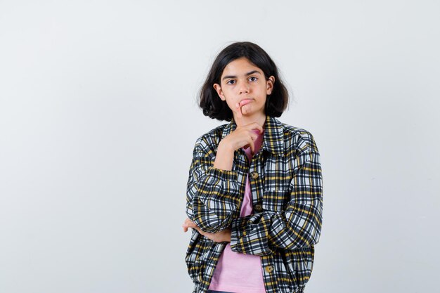 Young girl standing in thinking pose in checked shirt and pink t-shirt and looking pensive , front view.