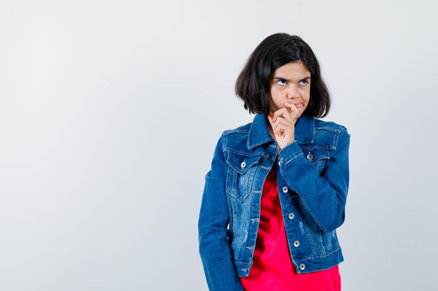 Young girl standing in thinking pose in red t-shirt and jean jacket and looking pensive , front view.