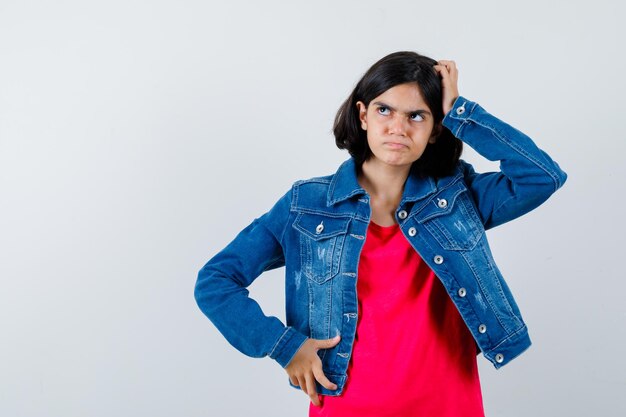 Young girl standing in thinking pose in red t-shirt and jean jacket and looking pensive , front view.