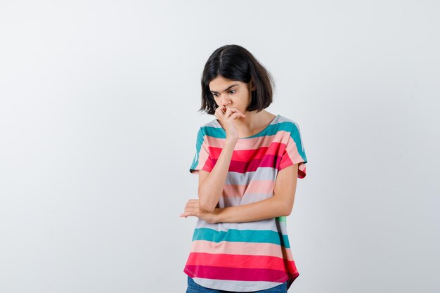 Young girl standing in thinking position, putting hand on mouth in colorful striped t-shirt and looking pensive , front view.