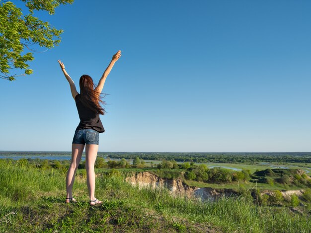 A young girl stands with her arms raised up to the sky. Relaxed young woman looking out into the view. Peaceful girl standing by a cliff enjoying the landscape. - outdoors. Full length portrait