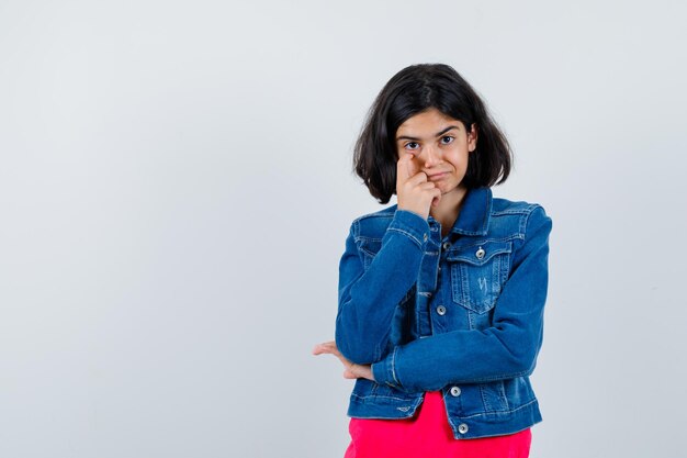 Young girl stretching eye area with index finger, smiling in red t-shirt and jean jacket and looking happy