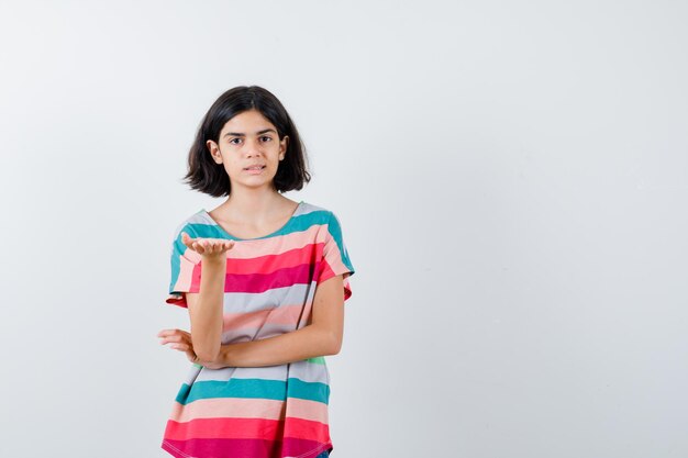 Young girl stretching hand as holding something in colorful striped t-shirt and looking serious. front view.