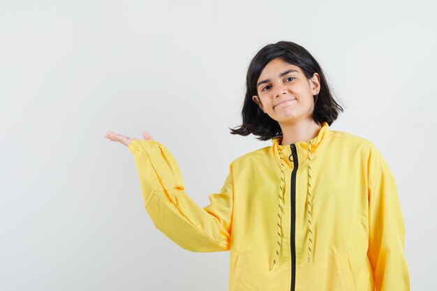 Young girl stretching hand left as holding something imaginary in yellow bomber jacket and looking serious