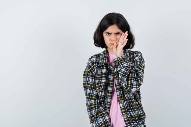 Giovane ragazza che allunga la mano vicino al viso, fa una smorfia in camicia a quadri e maglietta rosa e sembra arrabbiata. vista frontale.