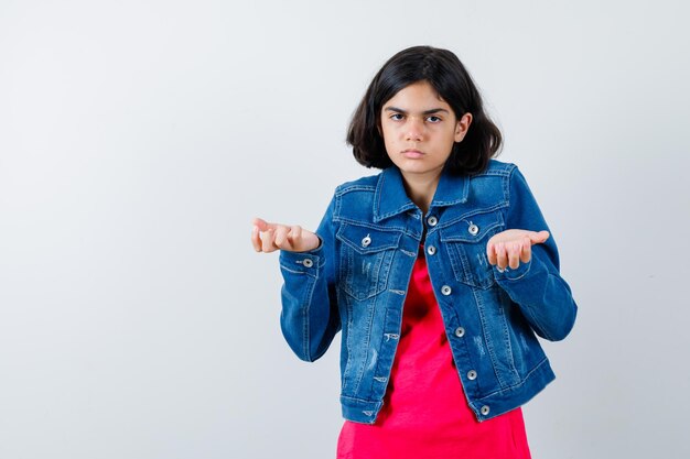 Young girl stretching hands in questioning manner in red t-shirt and jean jacket and looking baffled , front view.