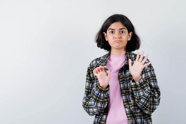 Young girl stretching hands to stop something in checked shirt and pink t-shirt and looking scared , front view.