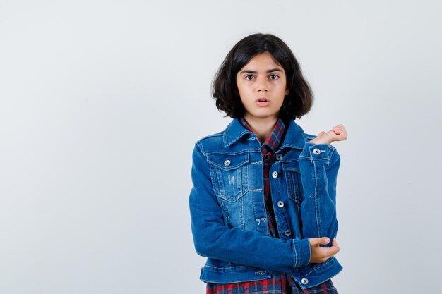 Young girl stretching one hand as holding something in checked shirt and jean jacket and looking serious. front view.