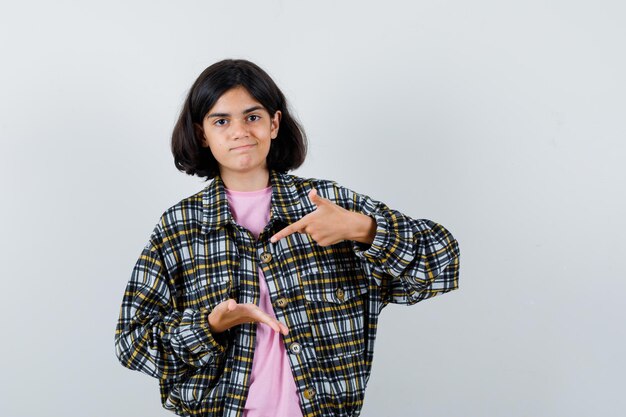 Giovane ragazza che allunga una mano mentre tiene qualcosa e lo indica con il dito indice in camicia a quadri e t-shirt rosa e sembra carina, vista frontale.