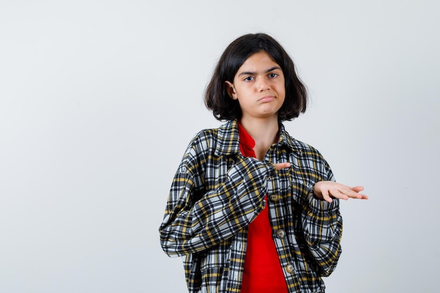 Giovane ragazza che allunga una mano mentre tiene qualcosa e lo indica con il dito indice in camicia a quadri e maglietta rossa e sembra seria. vista frontale.