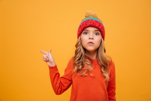 Young girl in sweater and hat pointing away and looking at the camera over orange