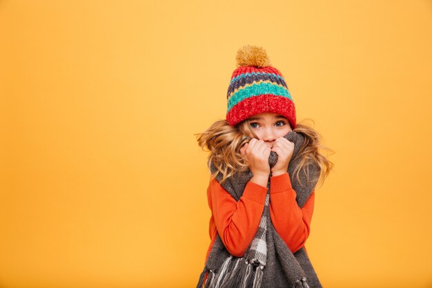 Young girl in sweater, scarf and hat having cold while looking at the camera over orange