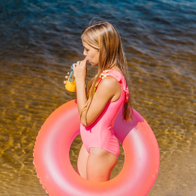 Молодая девушка в купальнике с плавательным кольцом пьет сок на пляже