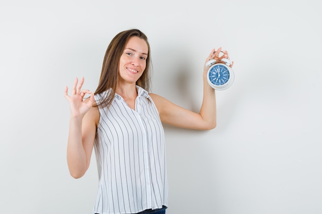 Young girl in t-shirt, jeans holding alarm clock with ok sign and looking cheerful , front view.