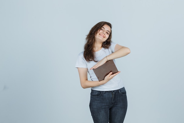 Young girl in t-shirt, jeans holding book while showing size sign and looking cheery , front view.